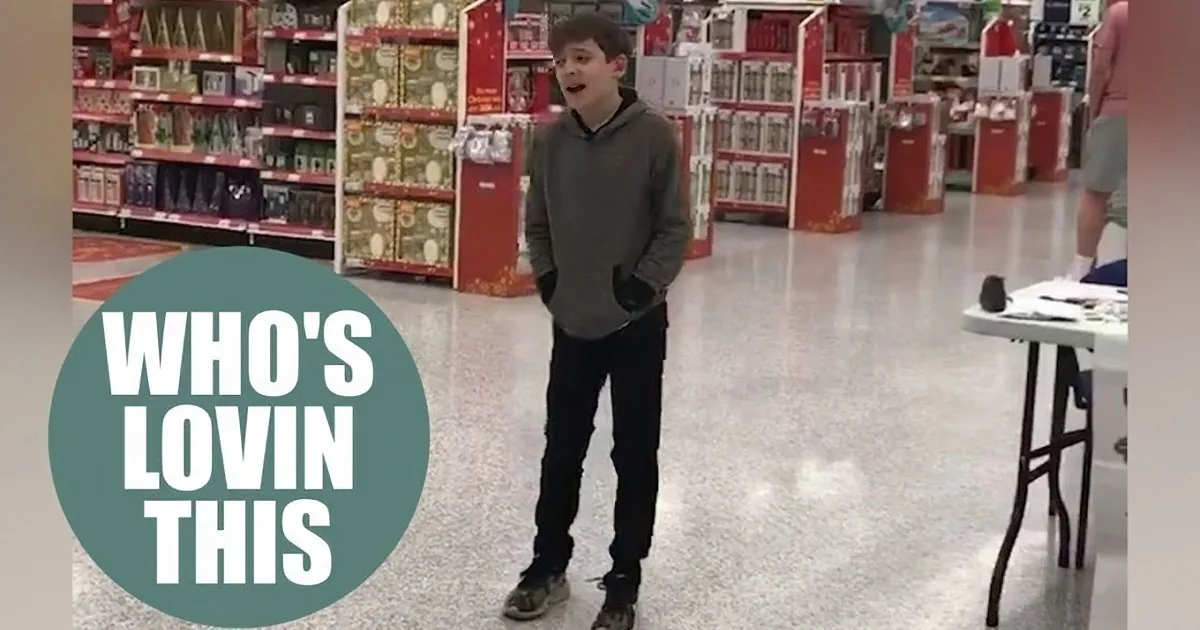 A boy stands in a store aisle, wearing a gray hoodie and black pants, with a surprised expression.