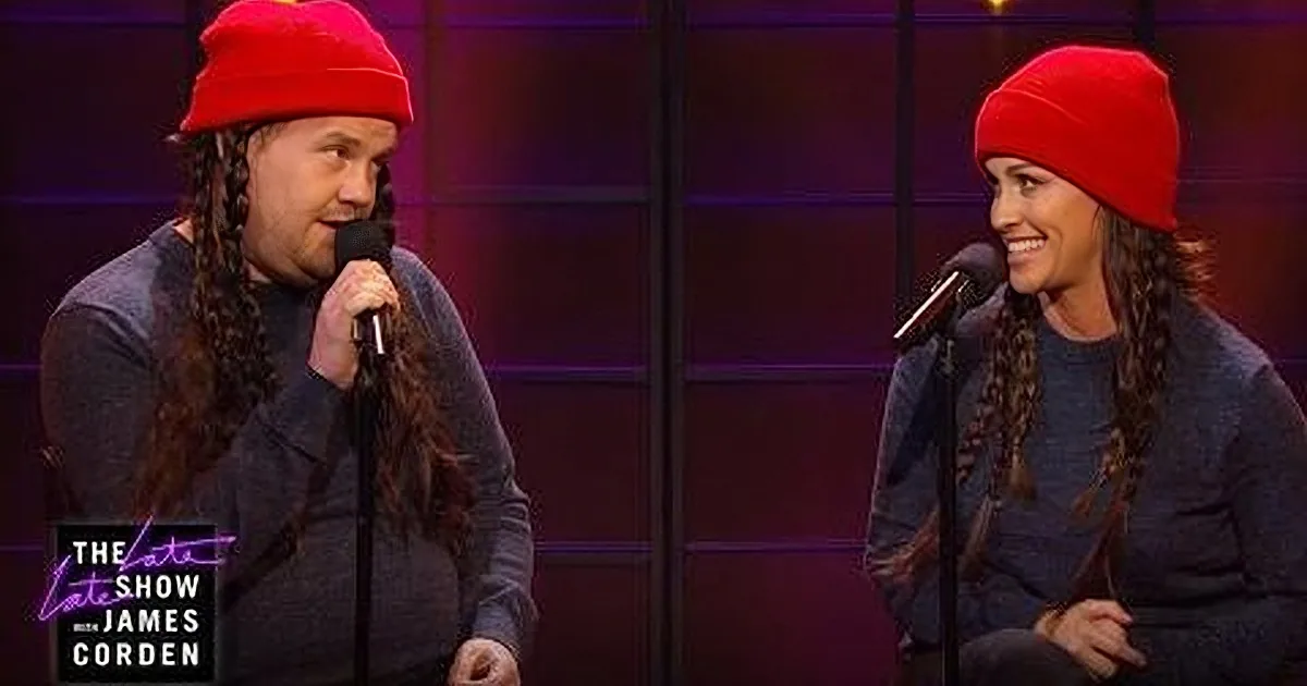 A man with long braids and a red beanie holds a microphone on stage, smiling at someone off-screen.