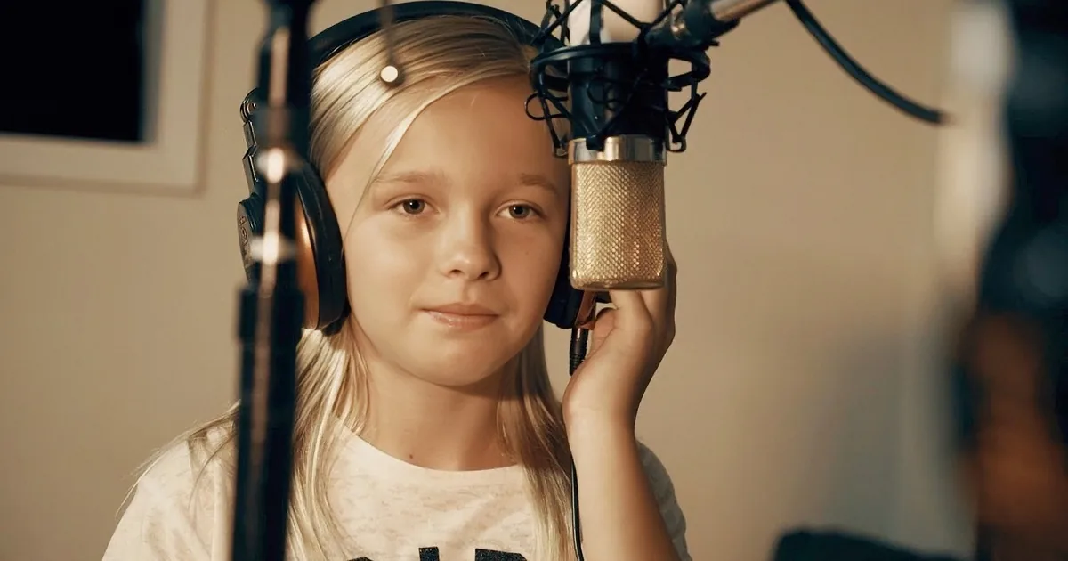 A young girl with blonde hair wearing headphones and holding a microphone in a recording studio.