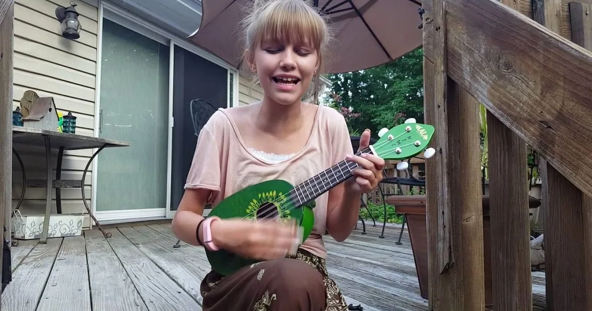 A girl plays a green ukulele on a wooden deck under an umbrella.