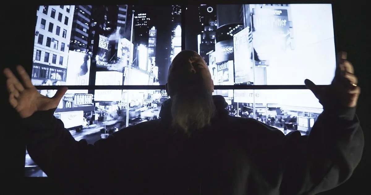 A person with long hair stands in front of a large screen displaying a cityscape at night.