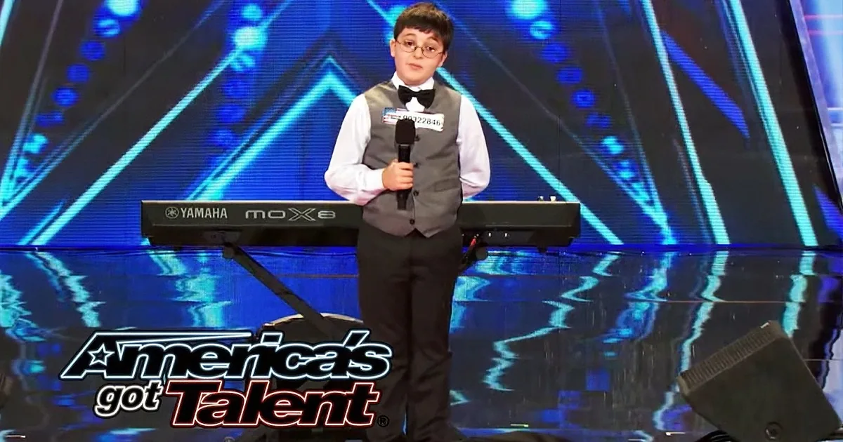 A young boy in a suit holds a microphone on stage with a Yamaha keyboard, performing on "America's Got Talent."