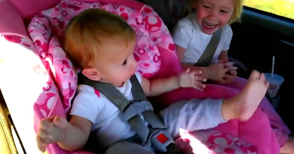 A baby in a pink car seat smiles while another child laughs in the backseat.