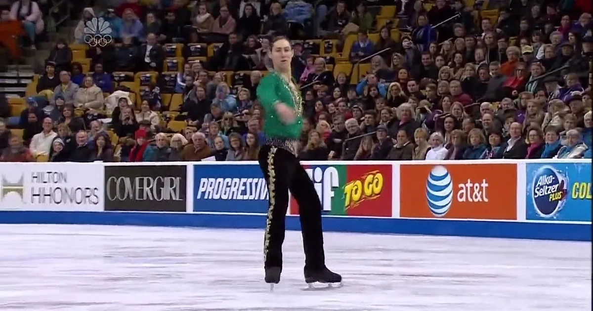 A figure skater in a green and black outfit performs a spin on the ice, surrounded by spectators and advertisements.
