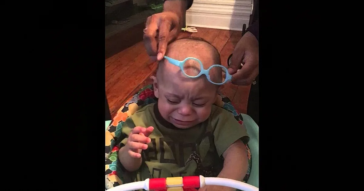 A baby in a high chair is having blue-framed glasses placed on their head by an adult.