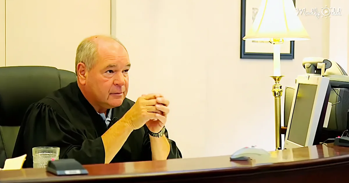 A judge in a black robe sits at a desk, hands clasped, with a computer and lamp in the background.