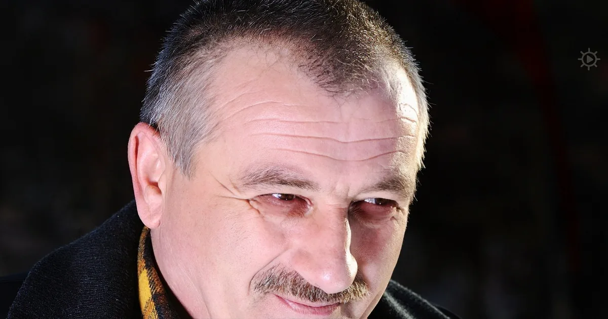 A man with short gray hair, a mustache, and a plaid shirt is looking down with a serious expression.