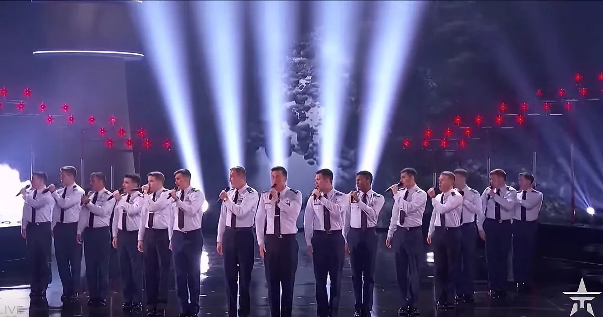 A group of men in white shirts and black pants stand in formation on a stage with bright lights and red spotlights.