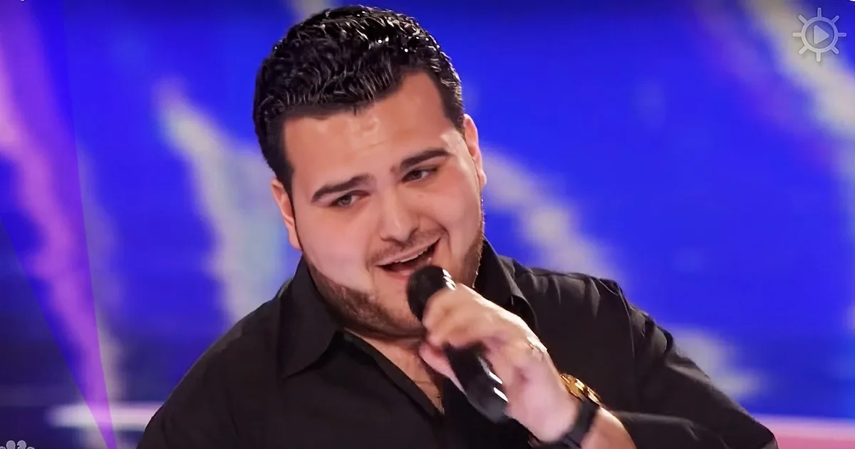 A man in a black shirt holds a microphone, smiling, with a blue and purple background.