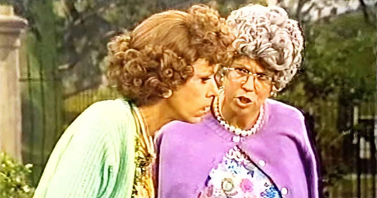 Two women, one in green and one in purple, appear to be engaged in a conversation outdoors.