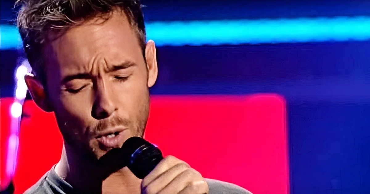 A man with a beard and short hair sings into a microphone, his eyes closed, against a red and blue background.