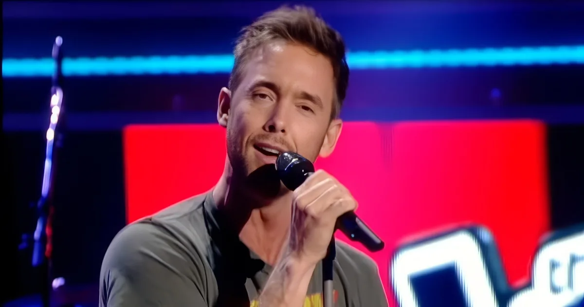 A man with short hair holds a microphone, singing on a stage with red and blue lighting.