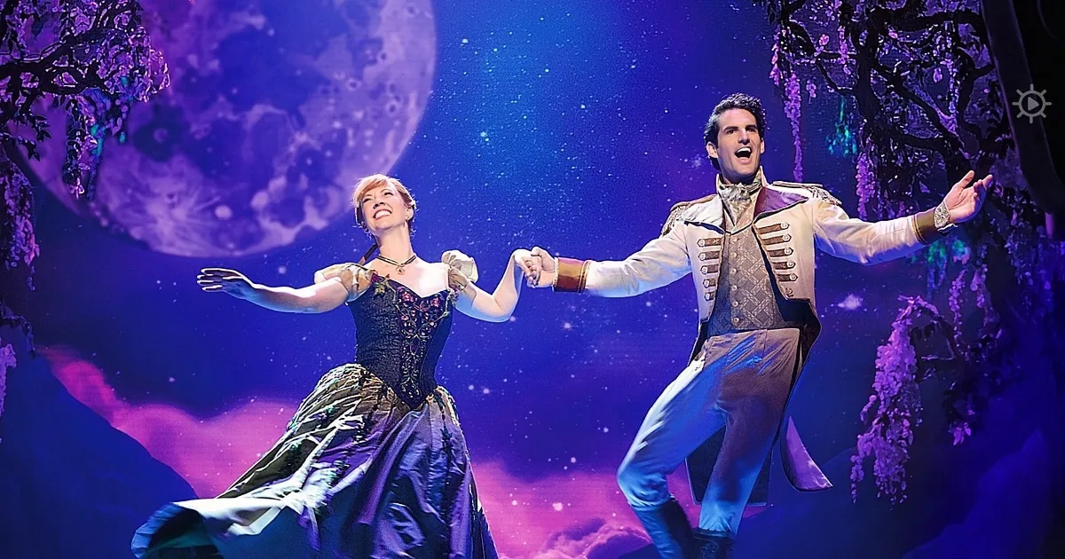 A couple in period costumes dances joyfully on stage under a starry night sky backdrop.