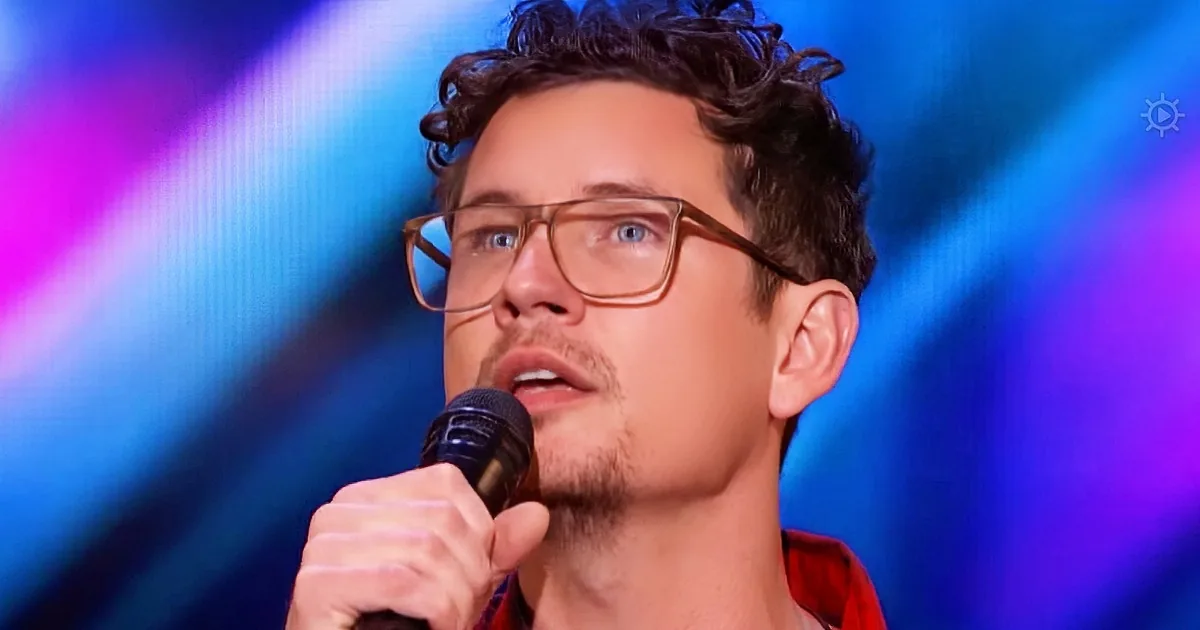A man with curly hair and glasses, wearing a red shirt, holds a microphone against a colorful, blurred background.