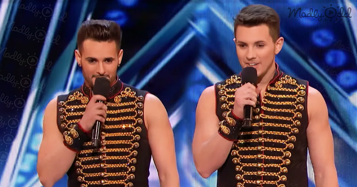 Two men in ornate black and gold vests hold microphones on stage, one with a mustache and the other with short hair.