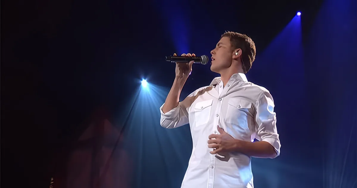 A man in a white shirt sings into a microphone on stage under blue spotlights.