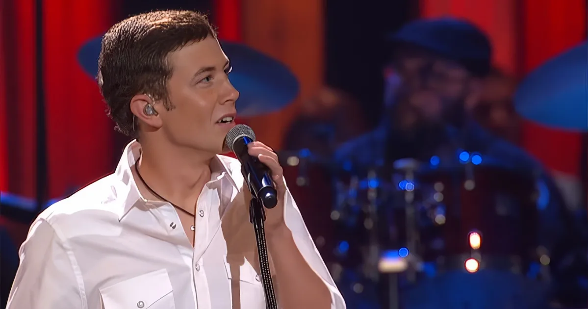 A man in a white shirt sings into a microphone on stage, with a drummer in the background.
