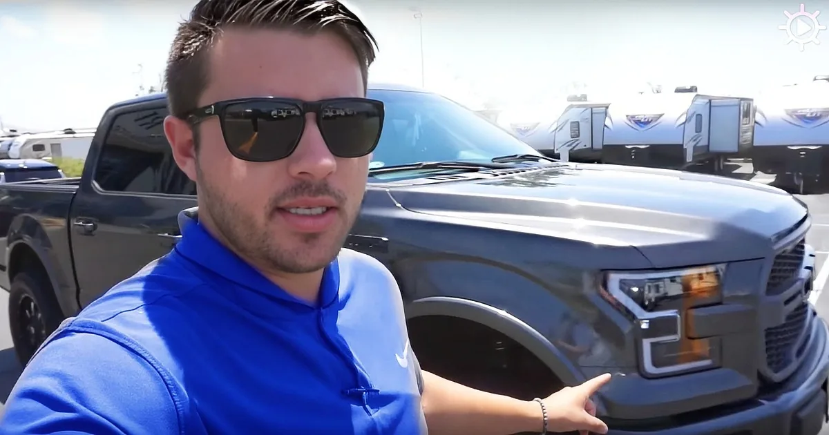 A man in a blue shirt and sunglasses stands in front of a gray pickup truck, gesturing with his hand.