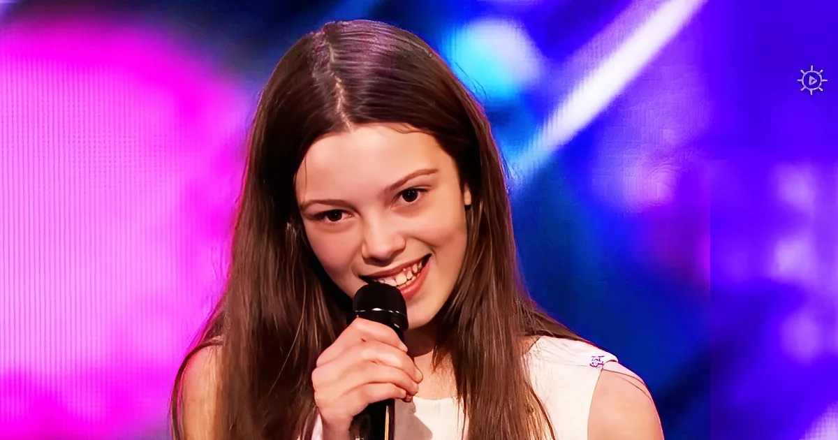 A young girl with long brown hair smiles while holding a microphone on a stage with colorful lights.