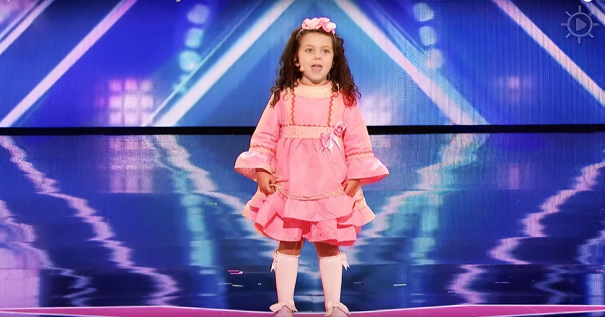 A young girl in a pink dress stands on stage with a colorful background.