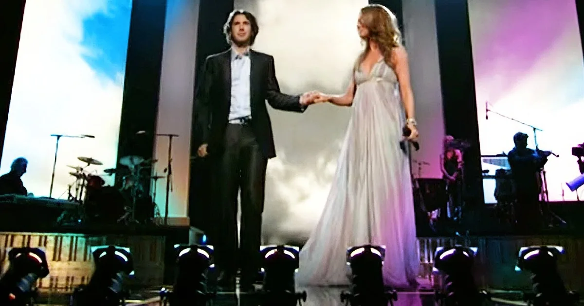 A man in a suit and a woman in a white dress are holding hands on a stage with a band in the background.