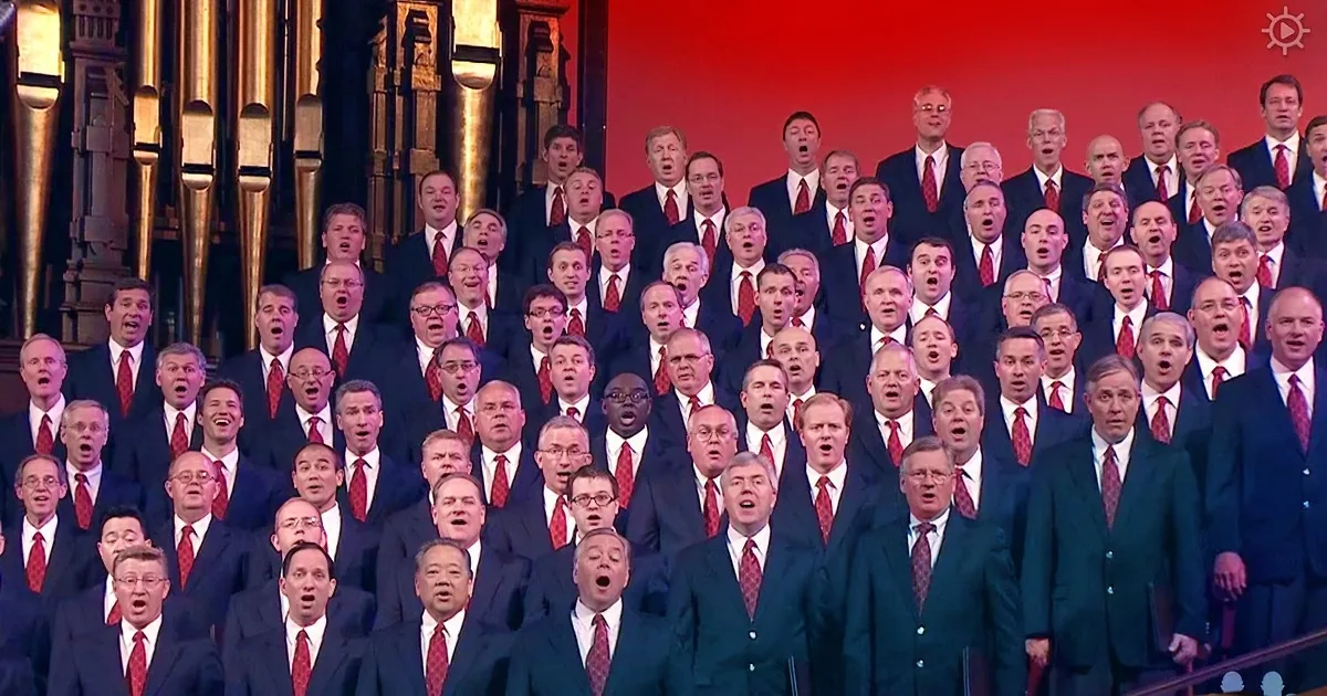 A large choir in dark suits and red ties sings together in a grand hall.