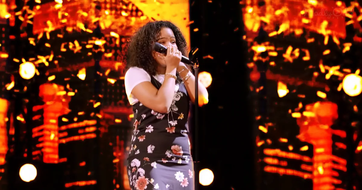A woman sings into a microphone on stage, wearing a floral dress, with a vibrant orange and black background.