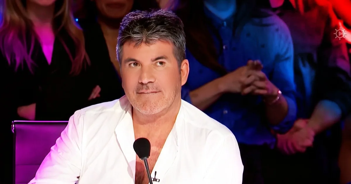 A man in a white shirt sits at a desk with a microphone, surrounded by a colorful audience.