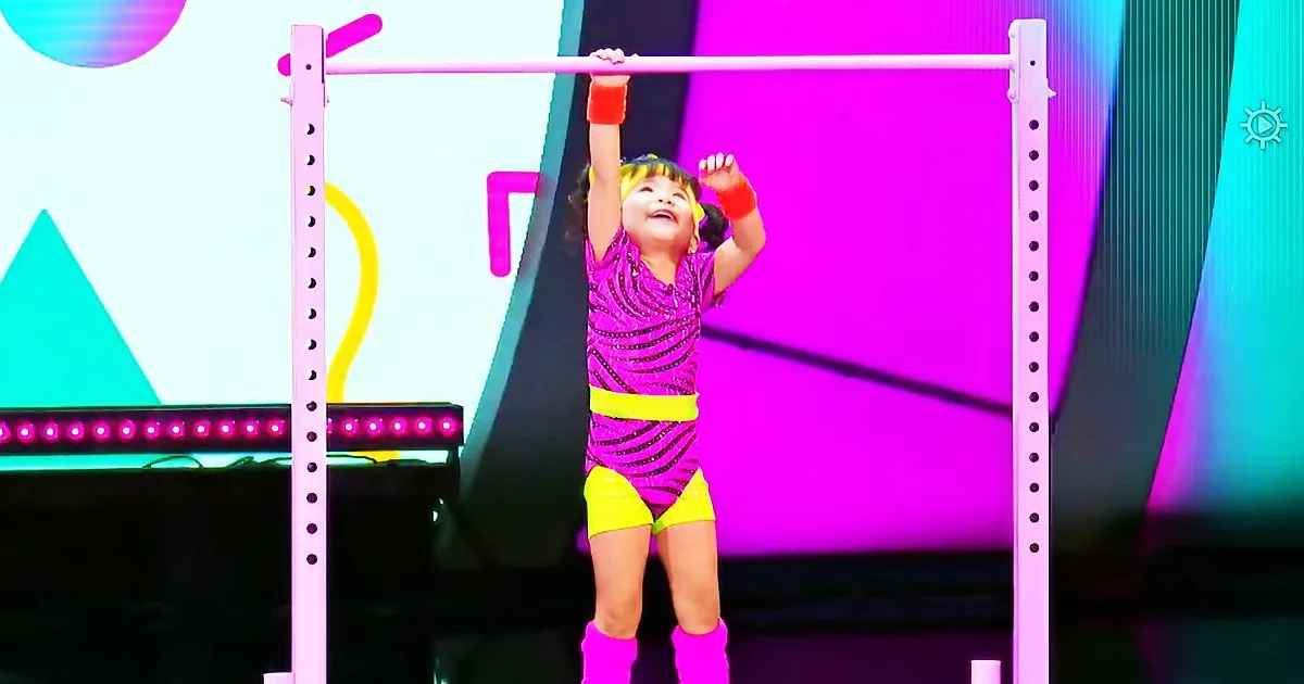 A child in bright pink and yellow outfit is hanging from a bar, smiling, with colorful stage lights in the background.