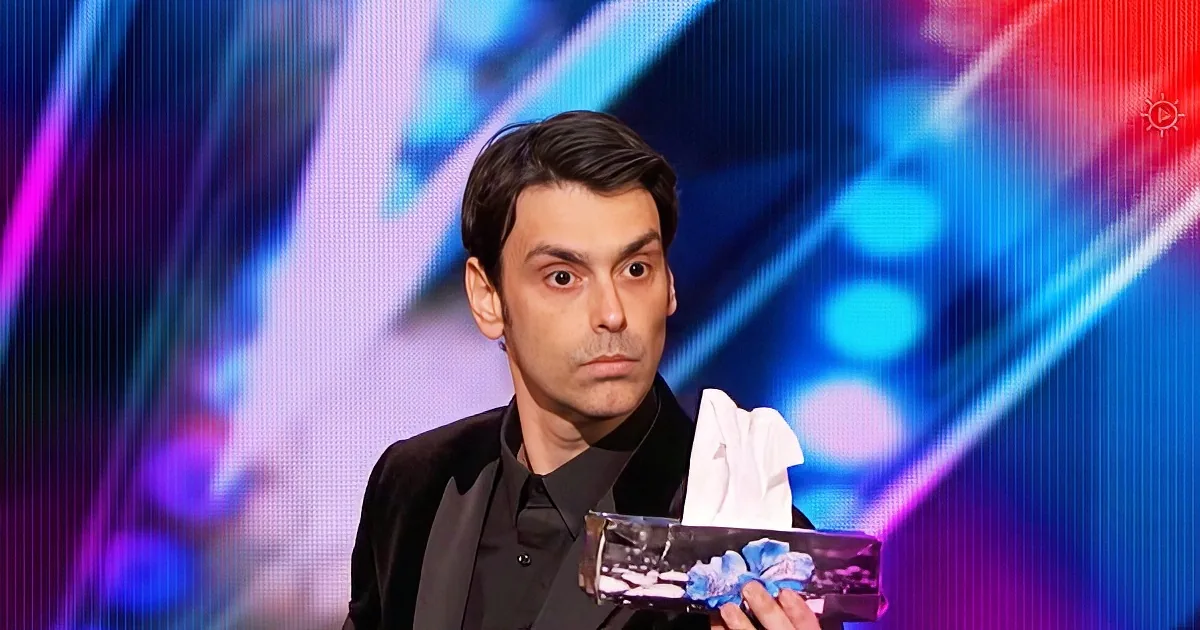 A man in a black suit holds a clear award with blue flowers, against a colorful, blurred background.