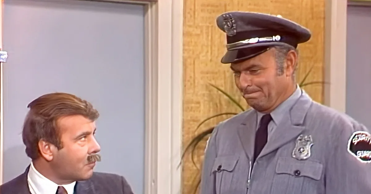 A man in a police uniform is smiling and talking to another man in a suit with a mustache.
