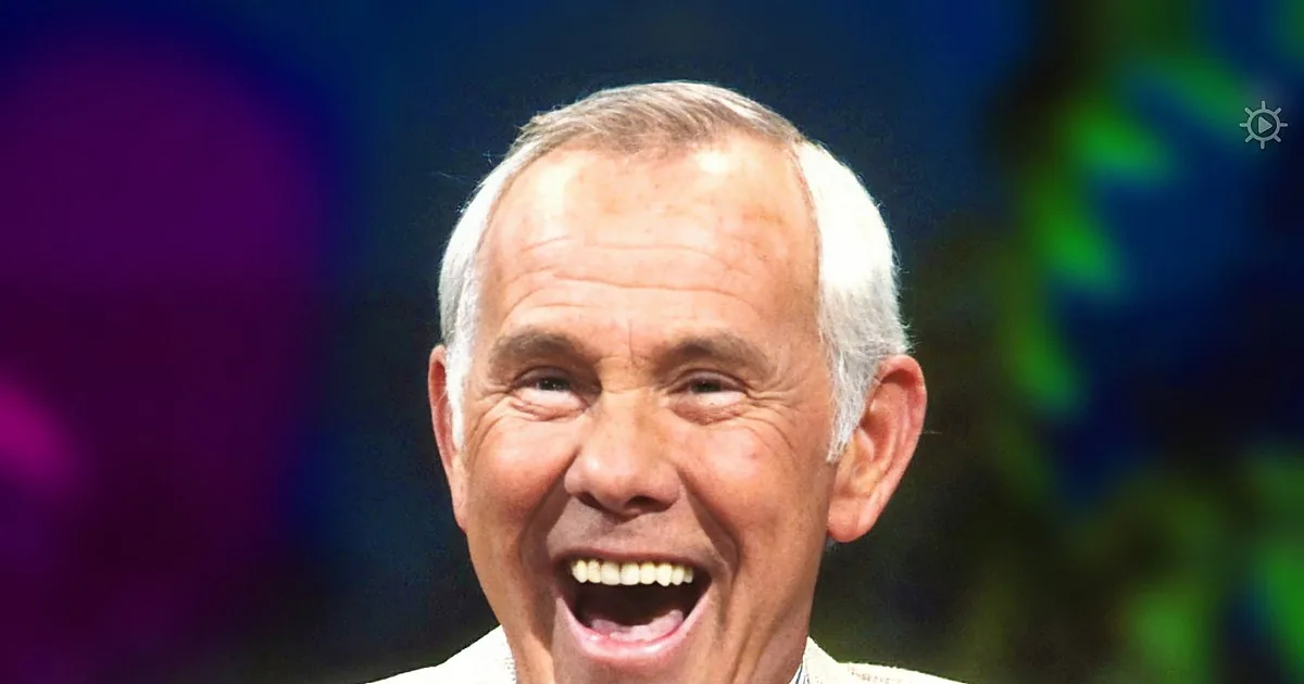 An older man with white hair, wearing a light suit and striped shirt, is smiling widely with his mouth open.