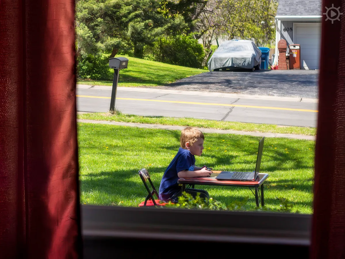 A boy sits at a small table outdoors, using a laptop, with a suburban street and house in the background.