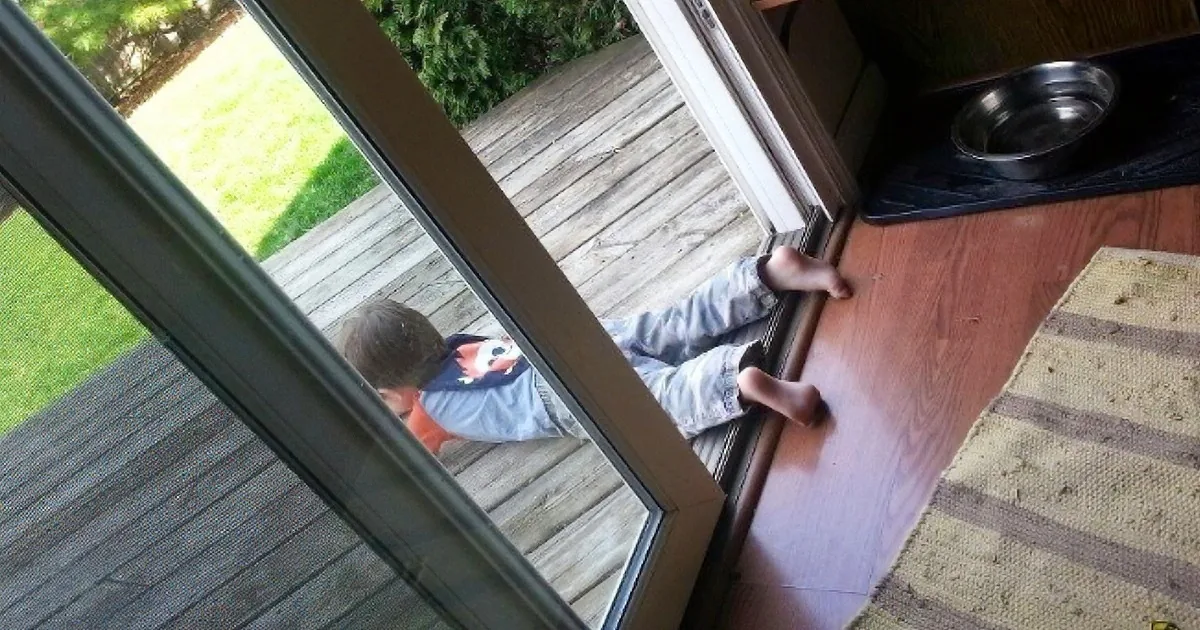 A child in a blue shirt and jeans is lying on a wooden deck, reaching for a door handle.