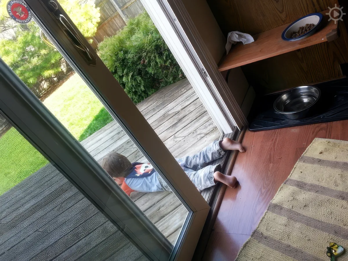 A child's legs are seen through a glass door, stepping onto a wooden deck.