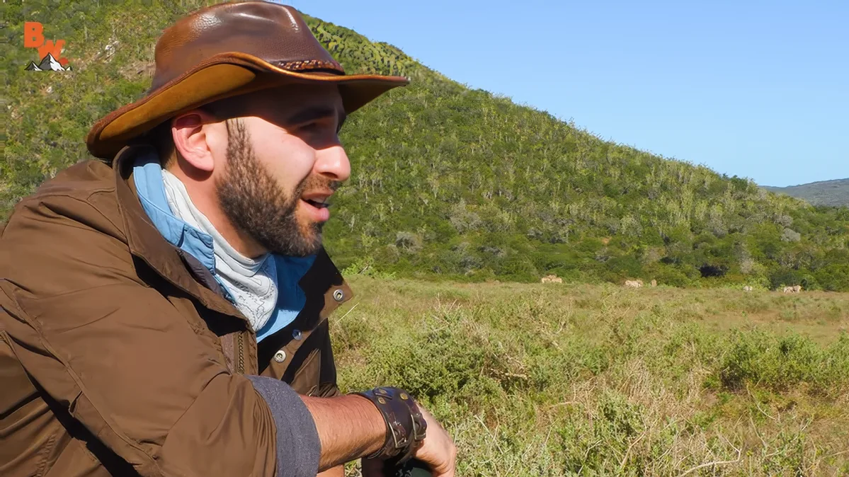 A man wearing a brown hat and jacket stands outdoors, surrounded by greenery and hills, with a clear blue sky in the...