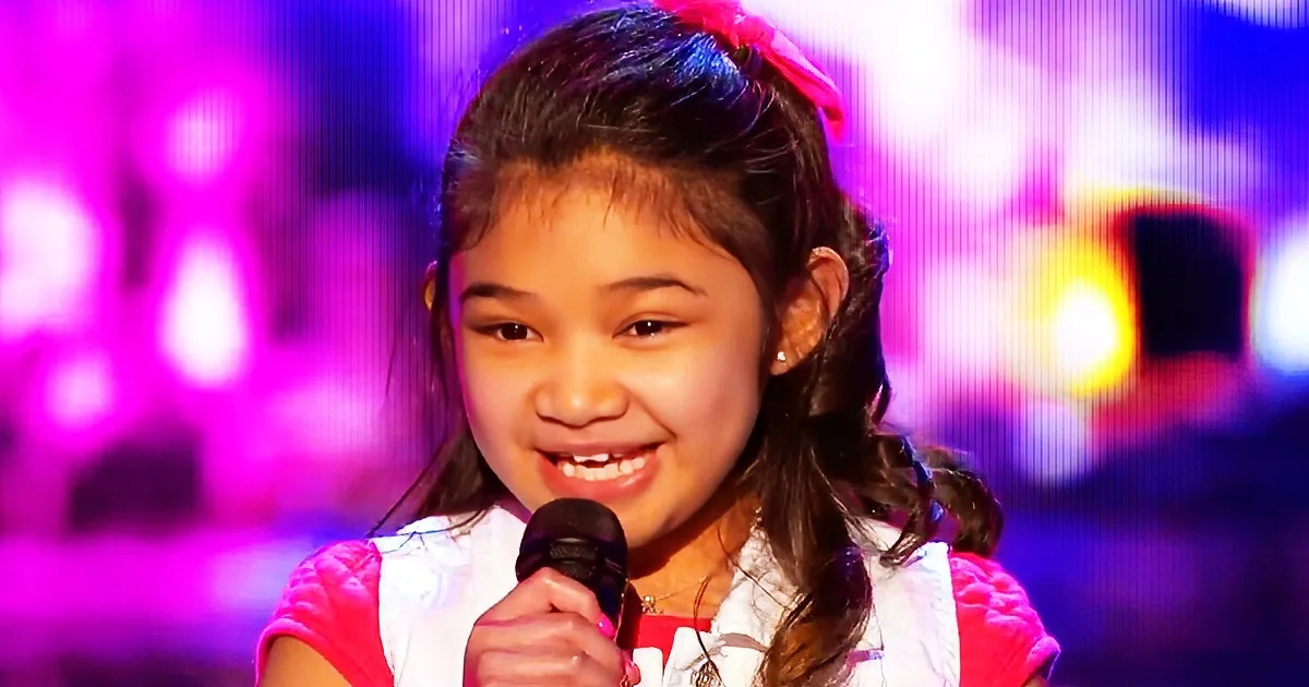 A young girl with a pink bow in her hair holds a microphone, smiling, against a vibrant purple and blue background.