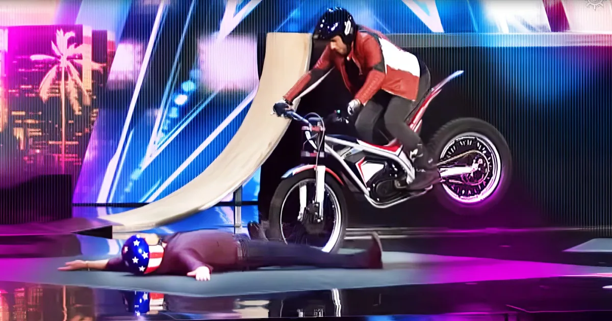 A motorcyclist performs a stunt on a ramp, wearing protective gear, with a cityscape backdrop.