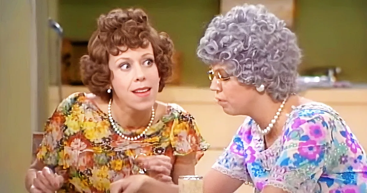 Two women in floral dresses, one with pearls, sit at a table, engaged in conversation.