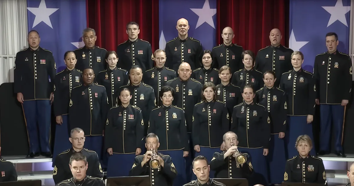 A military choir in uniform stands in formation, with a red curtain and stars in the background.