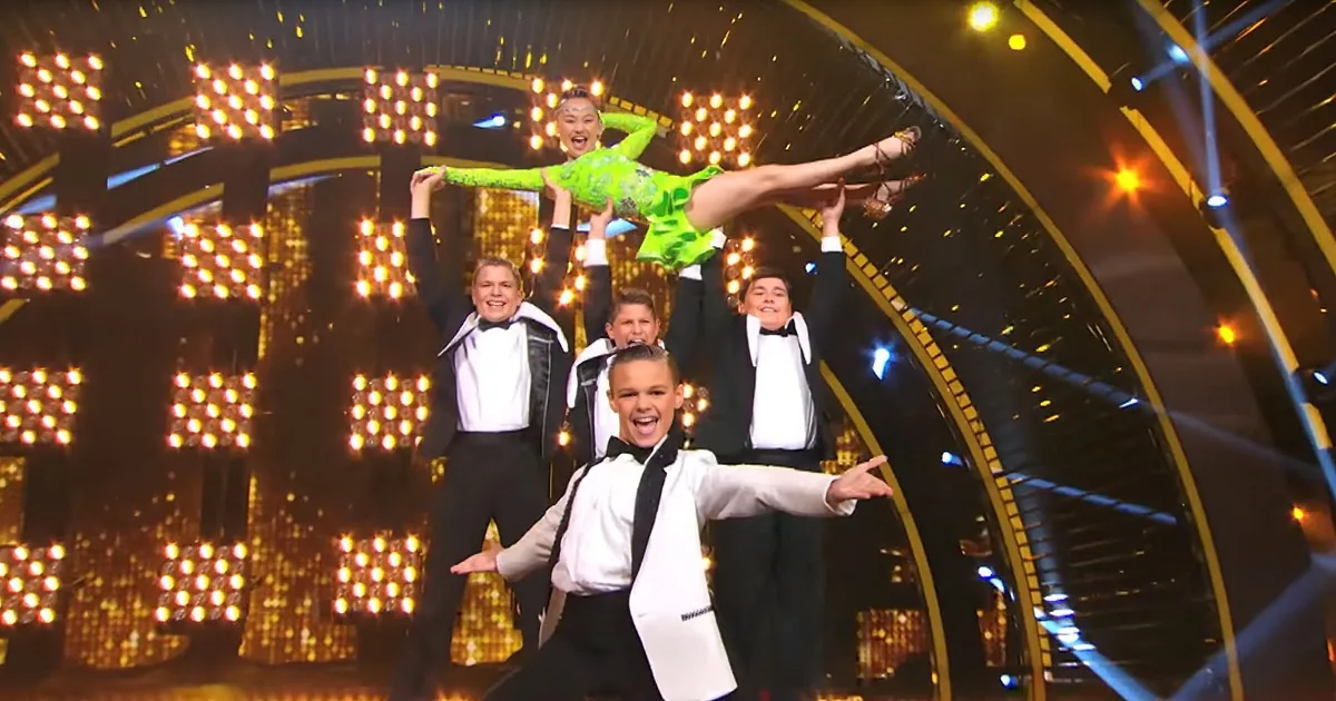 A performer in a green outfit is being lifted by four individuals in tuxedos on a stage with bright lights.
