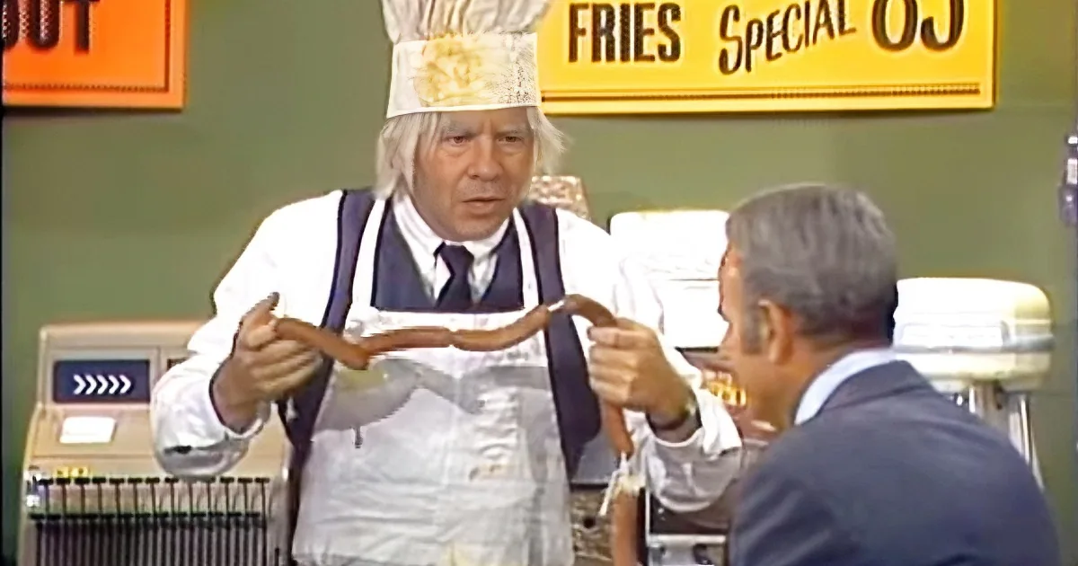 A man in a chef's hat and apron holds a sausage, while another person sits at a diner counter.