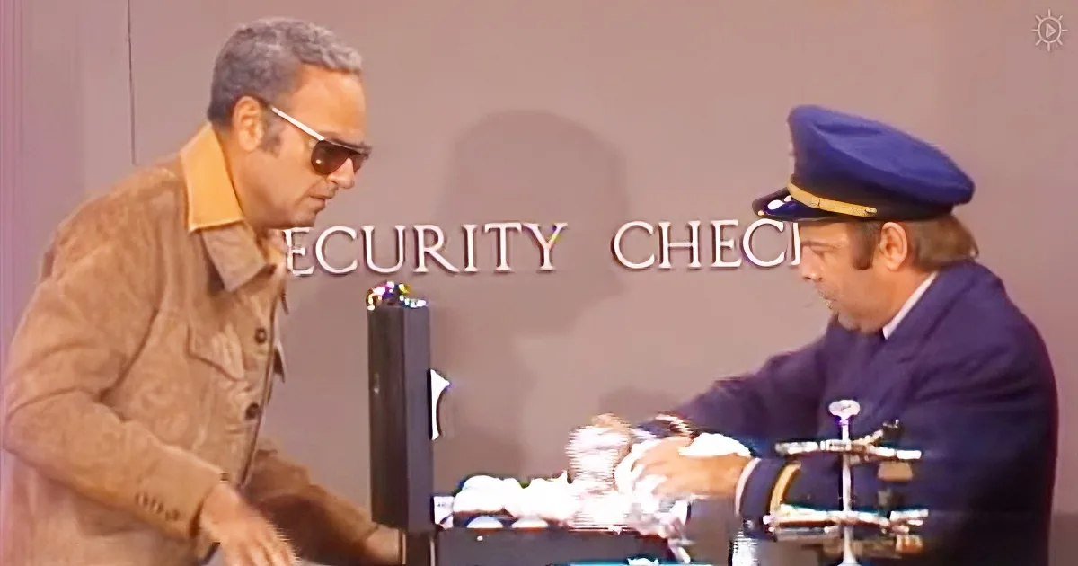 A man in sunglasses interacts with a security checkpoint officer at a desk.