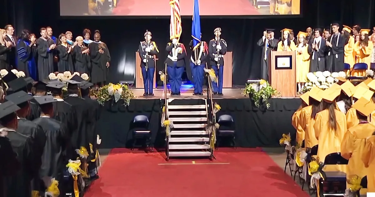 Graduates in caps and gowns stand on stage with flags, celebrating at a graduation ceremony.