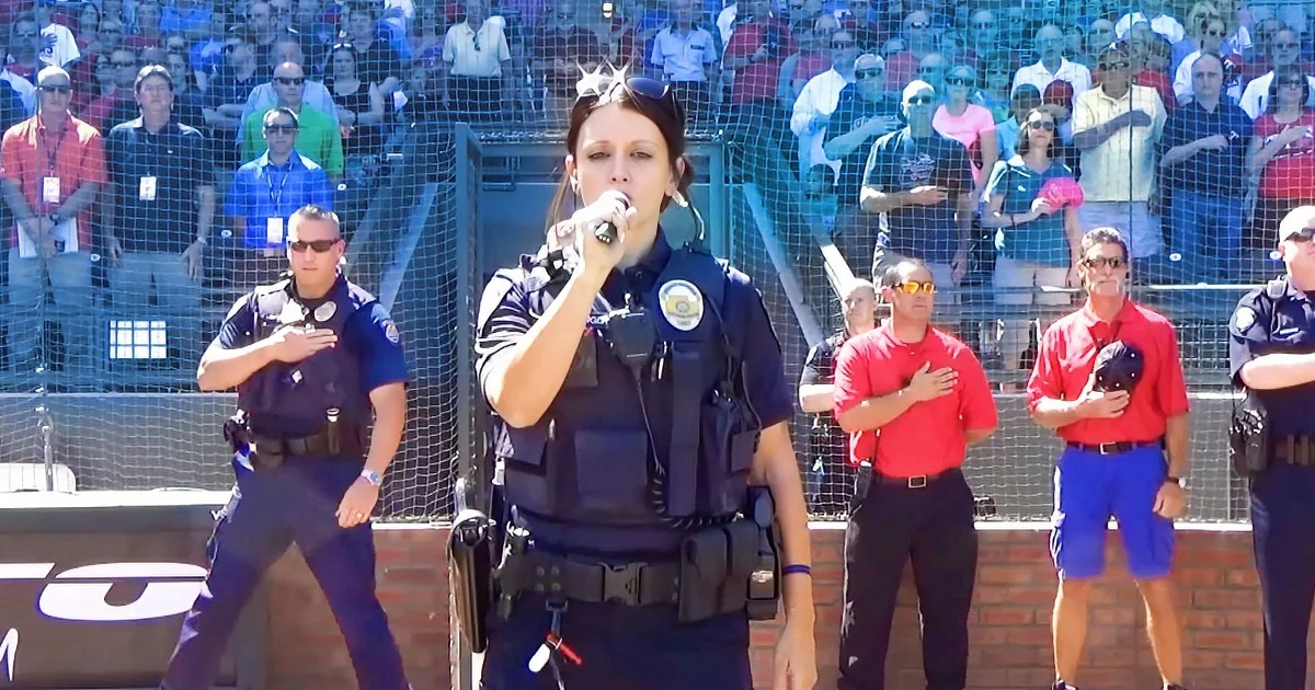 A police officer sings into a microphone while others stand with hands over hearts in front of a crowd.