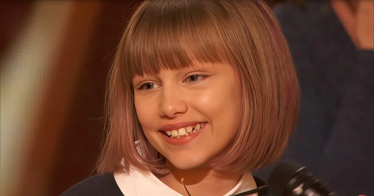 A young girl with short, light brown hair smiles while wearing a dark top, with a microphone visible in the foreground.