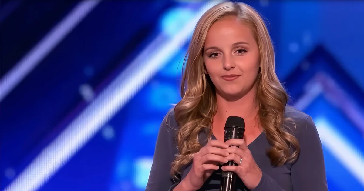 A woman with long blonde hair holds a microphone on a stage with a blue background.