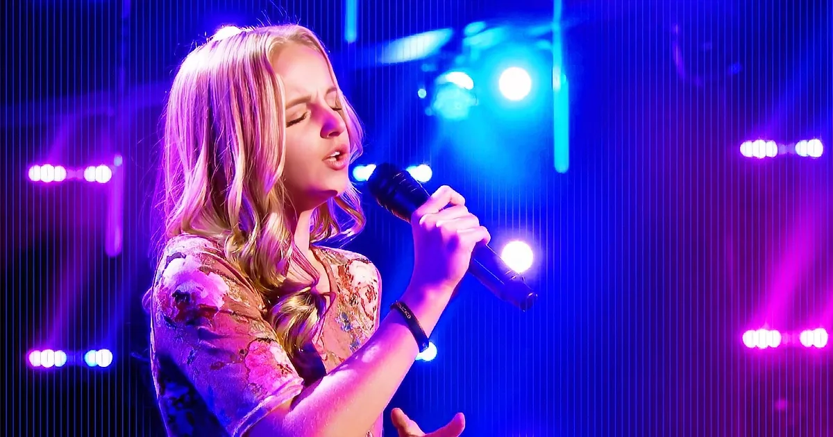A woman sings passionately into a microphone on stage under vibrant purple and blue lights.
