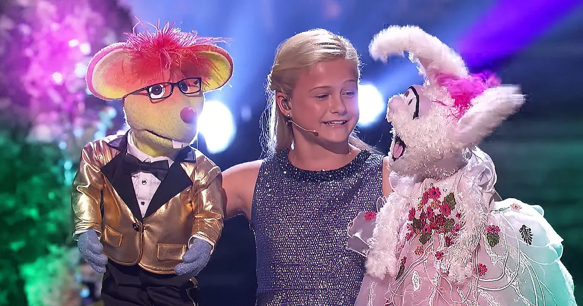 A young girl stands between two puppets, one in a gold suit and the other in a pink dress, on a stage with colorful lights.