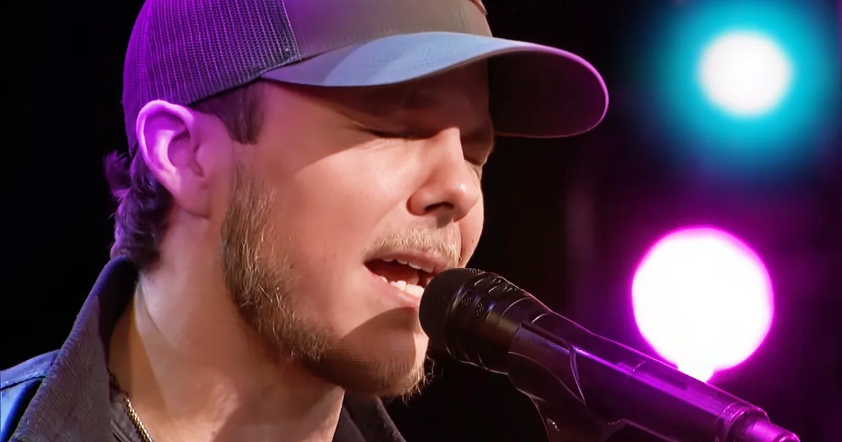 A person wearing a cap sings into a microphone with colorful stage lights in the background.